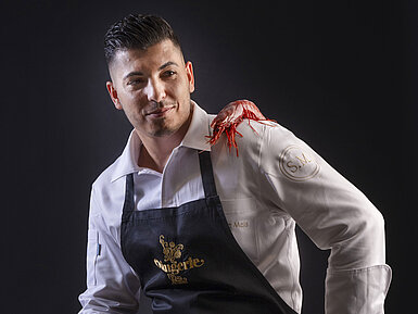 Küchenchef Simone Melis Küchenchef Simone Melis mit Schürze, einer roten Garnele auf der Schulter und einer Zitrone in der Hand.