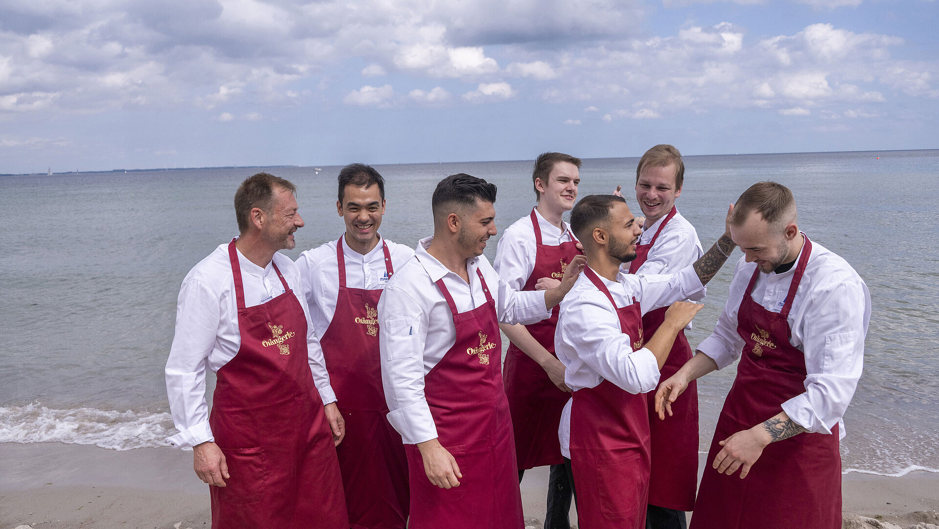 Team von der Orangerie Küchenteam der Orangerie im Maritim Hotel Timmendorfer Strand, am Strand in Kochuniformen und roten Schürzen stehend.