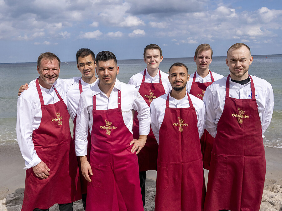 Team von der Orangerie Küchenteam der Orangerie im Maritim Hotel Timmendorfer Strand, am Strand in Kochuniformen und roten Schürzen stehend.