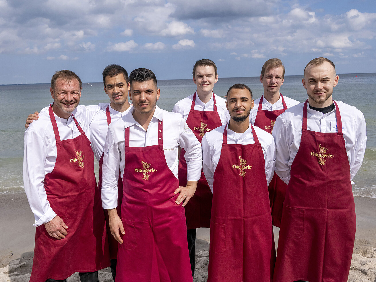Team von der Orangerie Küchenteam der Orangerie im Maritim Hotel Timmendorfer Strand, am Strand in Kochuniformen und roten Schürzen stehend.